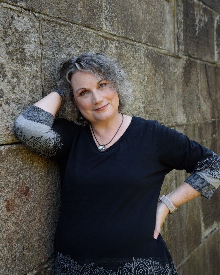 Photo of smiling woman with glasses and curly gray hair leaning casually against a stone wall.