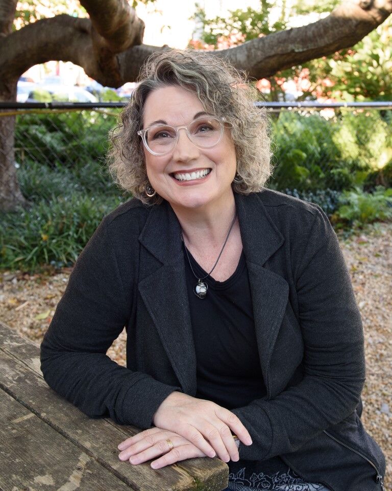 Photo of a grinning woman with glasses and curly gray hair seated at a bench in a park.
